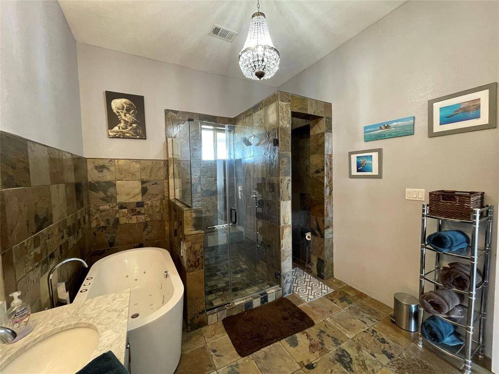 This bathroom features a luxurious spa-like ambiance with a jetted tub and a glass-enclosed shower. The slate tile flooring and walls add a natural touch, while a modern chandelier and framed art enhance the stylish decor. A metal shelving unit provides practical storage space.