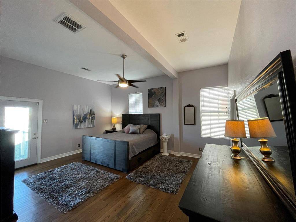 This spacious bedroom features hardwood floors, a large bed with storage, and a ceiling fan. It has a modern dresser with a TV and access to a balcony through a glass door. The room is well-lit with natural light and a stylish lamp.  Some furnishing's convey.