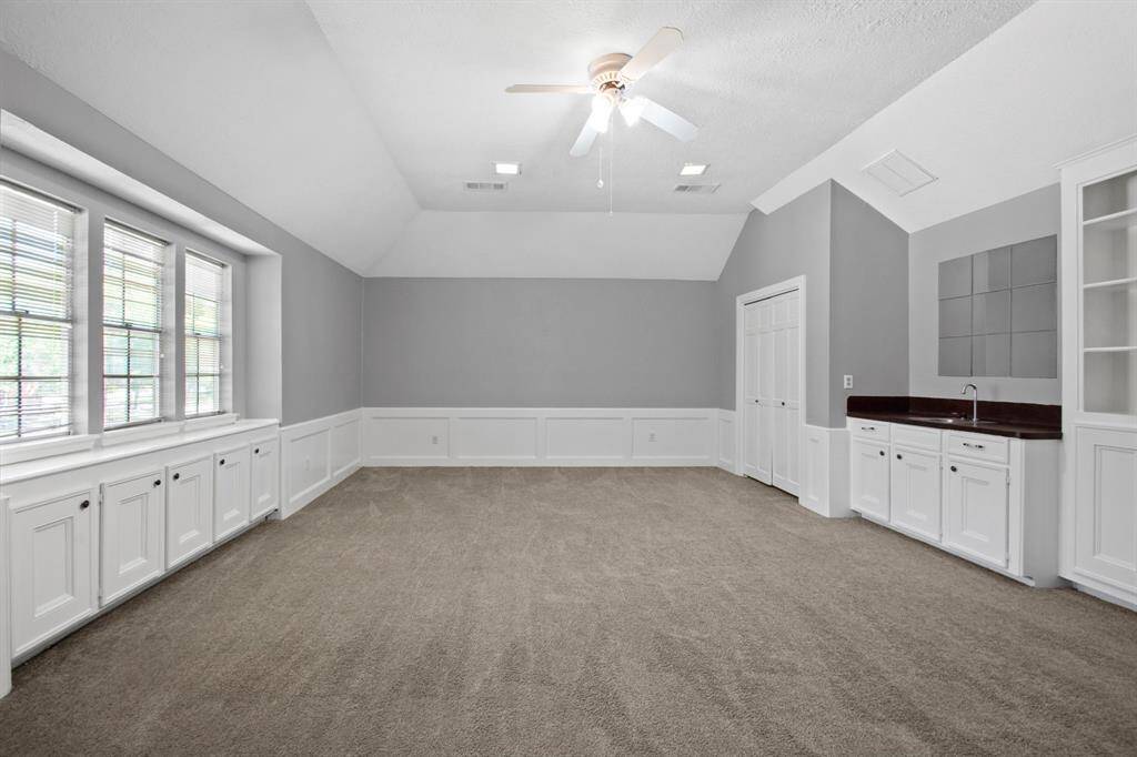 This spacious gameroom features neutral carpeting, a ceiling fan, and abundant natural light from large windows. It includes elegant wainscoting, a built-in wet bar with cabinetry, and versatile potential for a family or entertainment room.