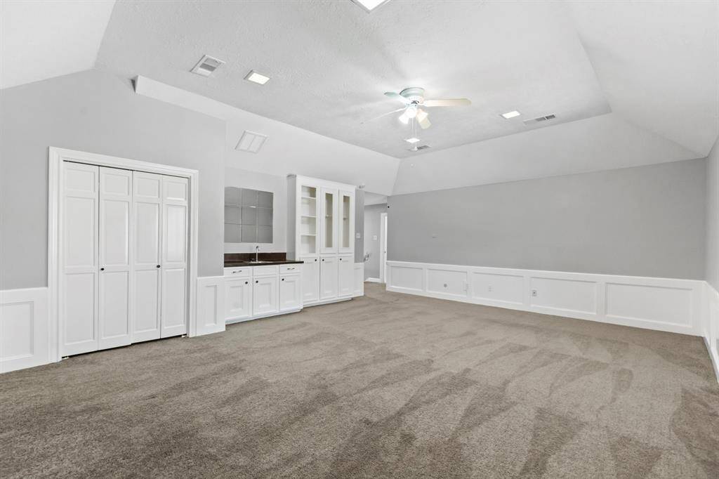 This spacious gameroom features a vaulted ceiling, neutral carpet, and light gray walls. It includes built-in cabinetry with a sink, a ceiling fan, and ample lighting, making it versatile for various uses.