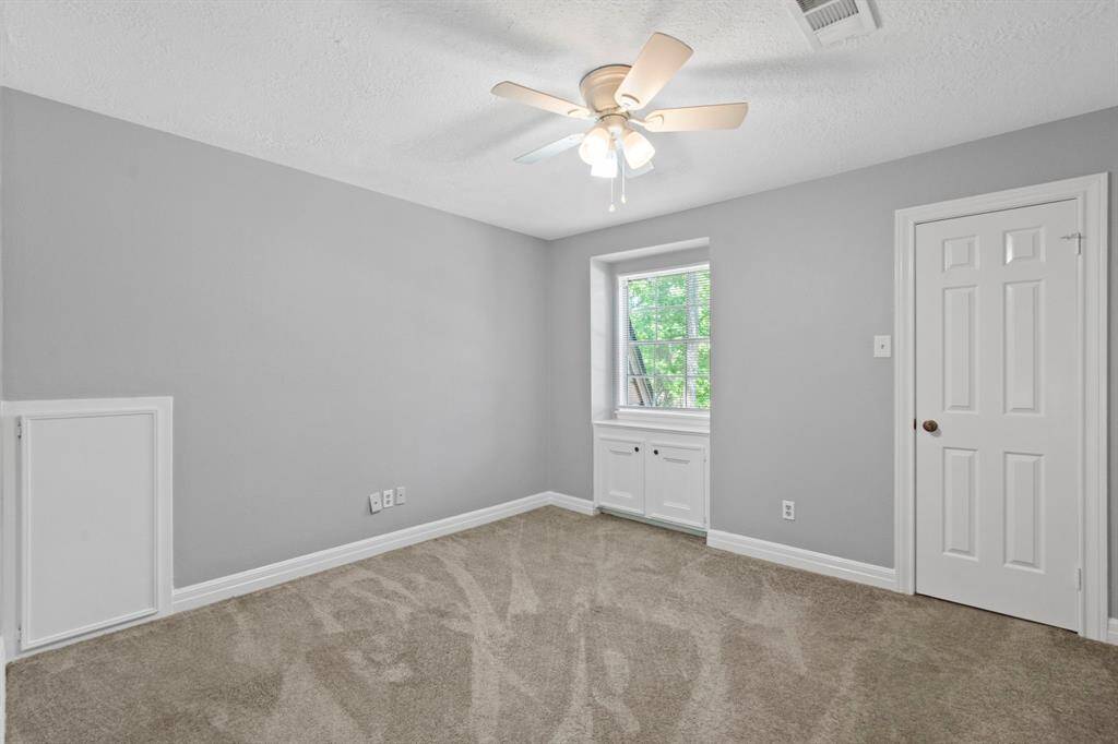 This bright room features soft gray walls, a plush carpet, and a ceiling fan for comfort. A window with built-in storage below lets in natural light, and there's a door providing easy access to an adjoining space.