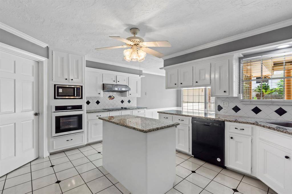 Bright, modern kitchen with white cabinetry, granite countertops, and a central island. Features tile flooring, built-in oven, microwave, and a ceiling fan. Large window provides natural light.