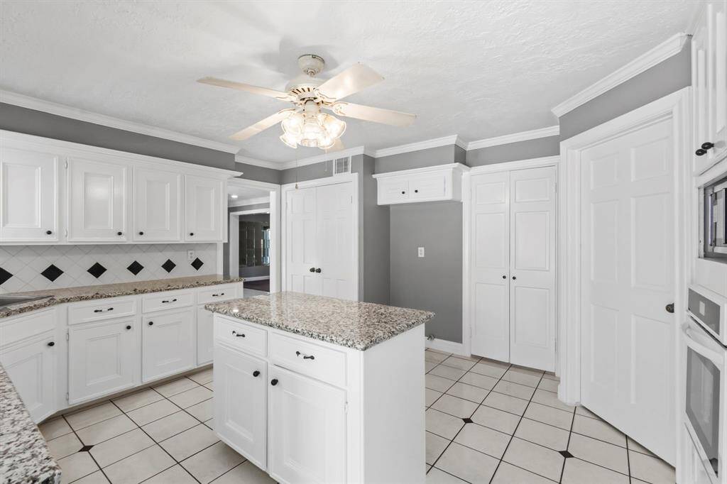 This kitchen features a modern design with white cabinetry, granite countertops, and a central island. It has a neutral color palette with gray walls and a tiled floor. A ceiling fan provides ventilation, and ample storage space is available.