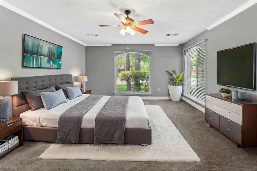 This spacious primary bedroom features a modern design with ample natural light from two large windows, neutral gray walls, and a ceiling fan.