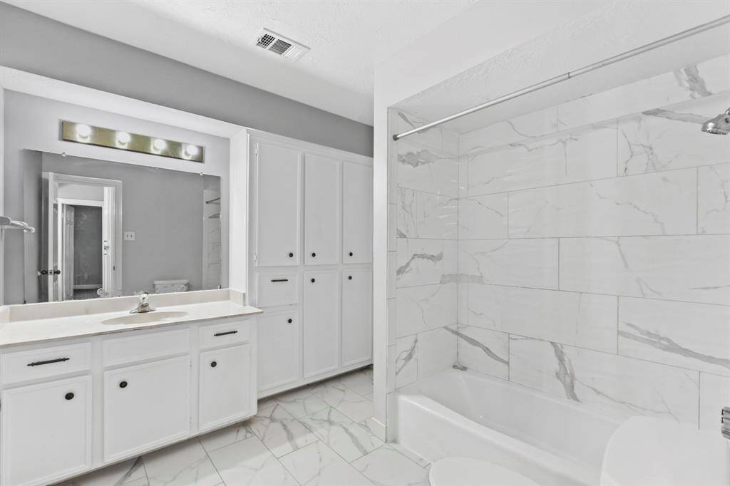 This bright bathroom features a sleek design with white cabinetry, ample storage, and elegant marble-patterned tiles. The large vanity mirror and modern fixtures enhance the spacious feel.