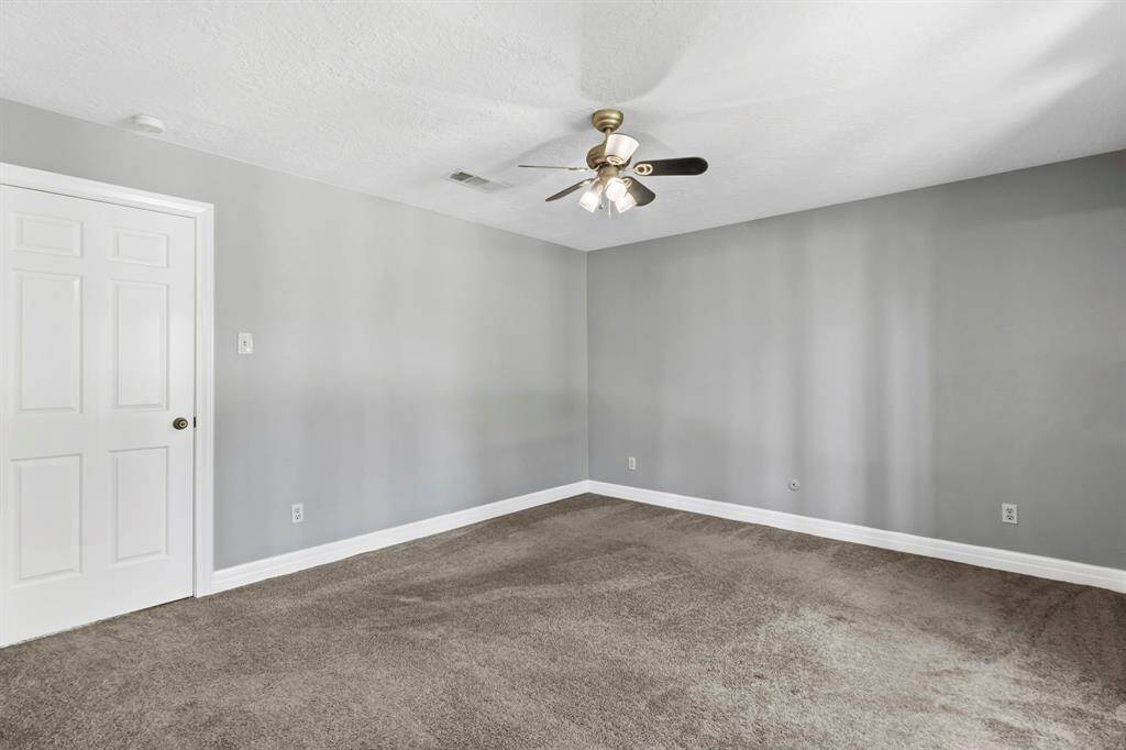 This room features a spacious layout with neutral gray walls and plush carpeting, complemented by a ceiling fan and ample natural light. Ideal for a bedroom or office space.