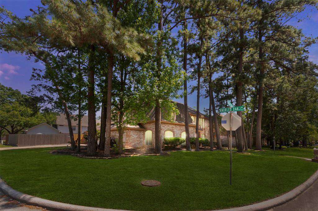 Charming brick home on a corner lot, surrounded by tall trees offering ample shade. The well-maintained lawn and quiet neighborhood setting create a serene atmosphere.