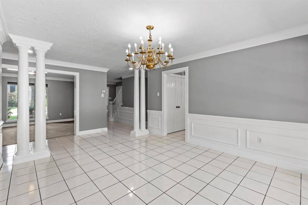 This elegant room features a spacious layout with white tile flooring, gray walls, and classic white wainscoting. It includes decorative columns and a gold chandelier, creating a sophisticated atmosphere. Perfect for formal dining or a stylish living area.