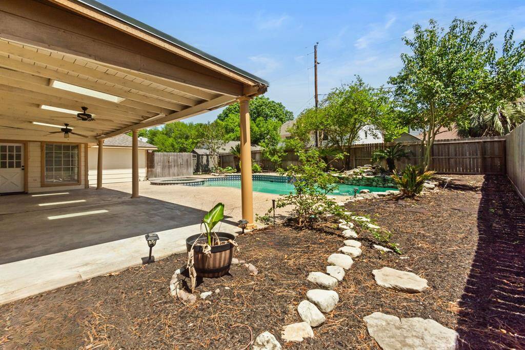 This backyard features a spacious covered patio with ceiling fans, perfect for outdoor gatherings. It overlooks a charming pool surrounded by landscaped greenery and a privacy fence, offering a serene retreat.