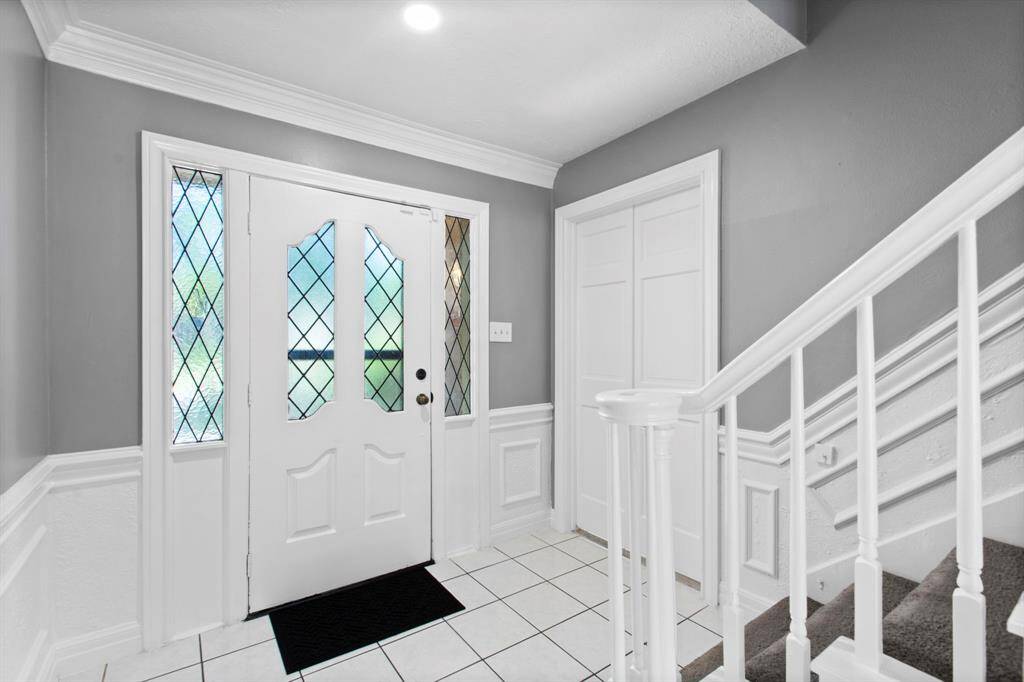 This entryway features a white front door with decorative glass panels, tiled flooring, and a staircase with white railing. The space has gray walls with white trim, creating a clean and welcoming atmosphere.