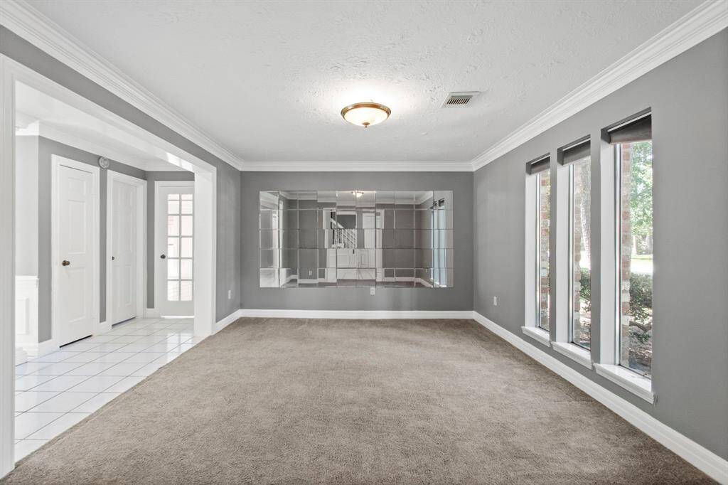 Bright, spacious room with large windows and a mirrored wall, featuring neutral gray walls and plush carpeting. The adjacent area with tiled flooring has access to additional rooms providing an open and airy feel.