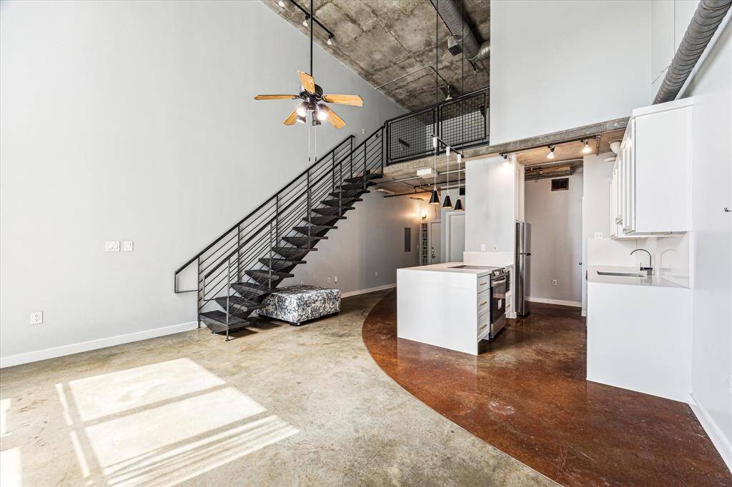 This modern loft features an open floor plan with high ceilings and industrial touches. The space includes a sleek kitchen island, metal staircase leading to a mezzanine, polished concrete floors, and large windows that let in ample natural light.