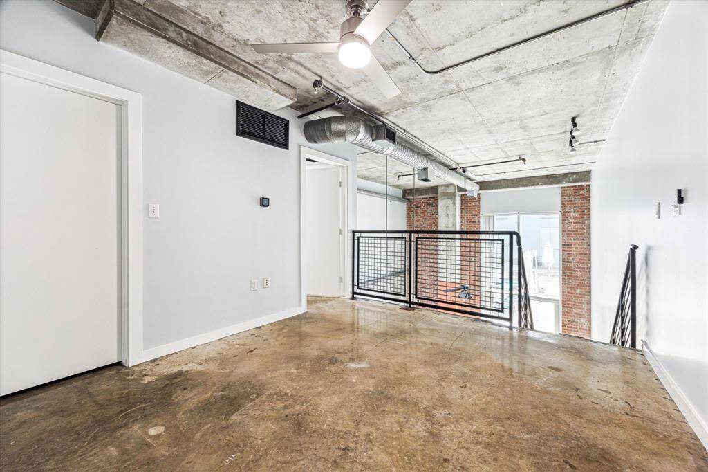 This space features an industrial-style loft with exposed concrete ceilings and ductwork. It has a modern, open layout with polished concrete floors, a ceiling fan, and brick accent wall. Large windows provide natural light, and a metal railing adds a sleek touch.