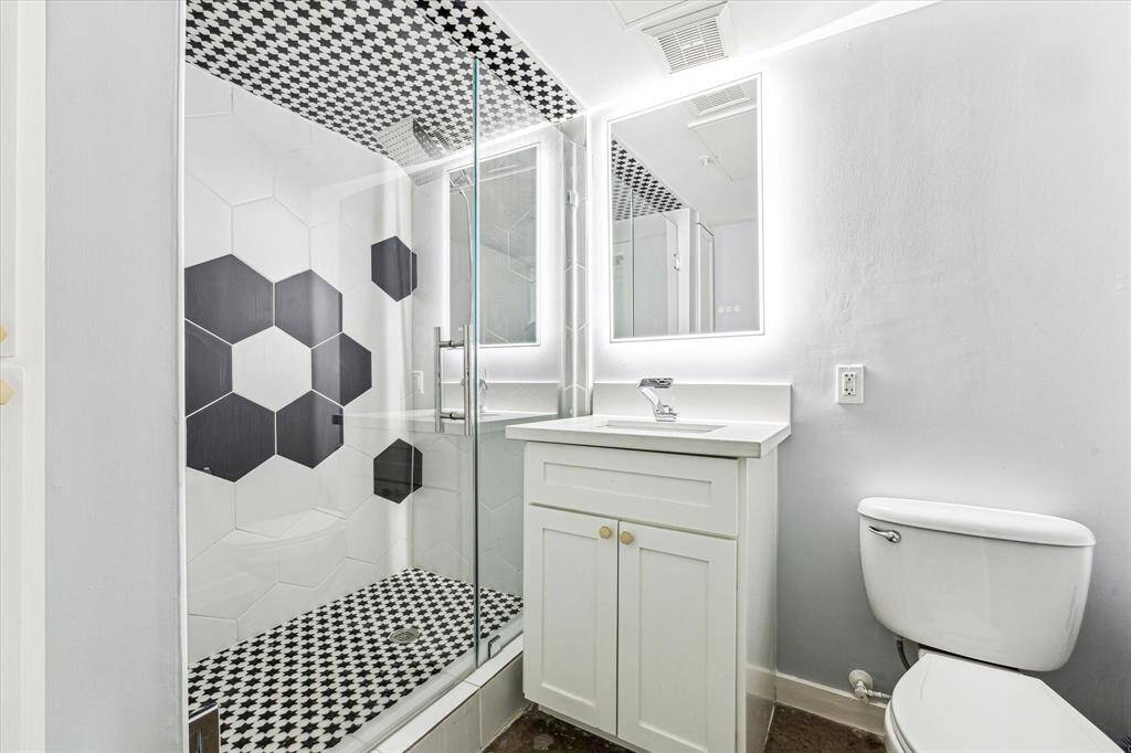 This modern bathroom features a sleek glass-enclosed shower with stylish black and white geometric tiles. The vanity includes an illuminated mirror and white cabinetry, complementing the clean and contemporary design.