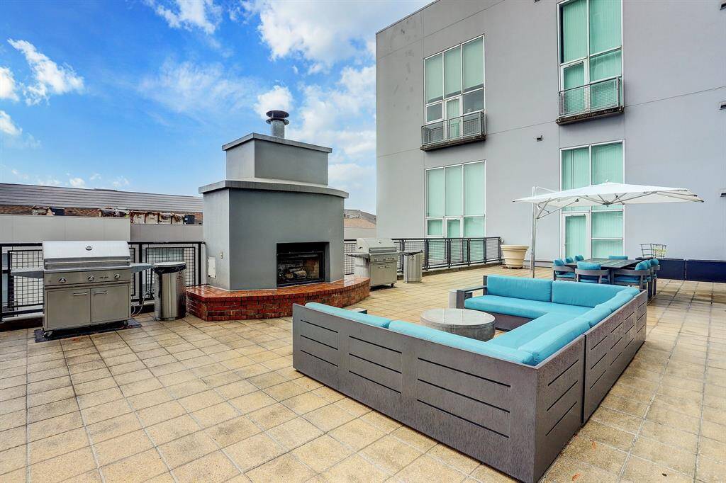 Modern outdoor terrace featuring a spacious seating area with turquoise cushions, a large fireplace, and two gas grills. Ideal for entertaining, it includes a dining table with an umbrella, set against a backdrop of contemporary architecture.