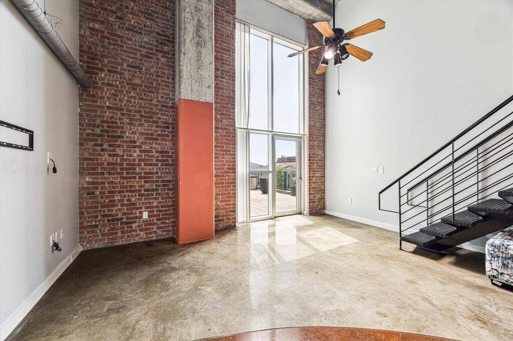 This spacious loft features high ceilings, exposed brick walls, and large floor-to-ceiling windows offering ample natural light. The open layout includes a modern industrial staircase leading to an upper level, perfect for versatile living arrangements.