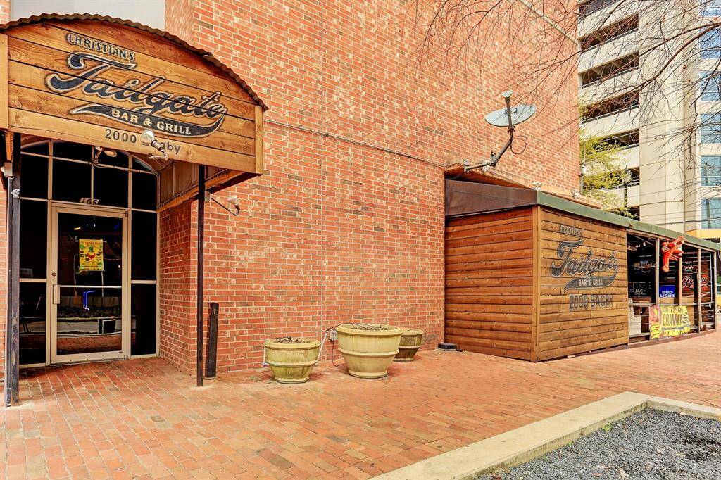 This photo shows a rustic bar and grill with a brick exterior, located in a lively urban area. The entrance is inviting with large planters and a wooden sign. Ideal for those seeking local dining and entertainment options nearby.