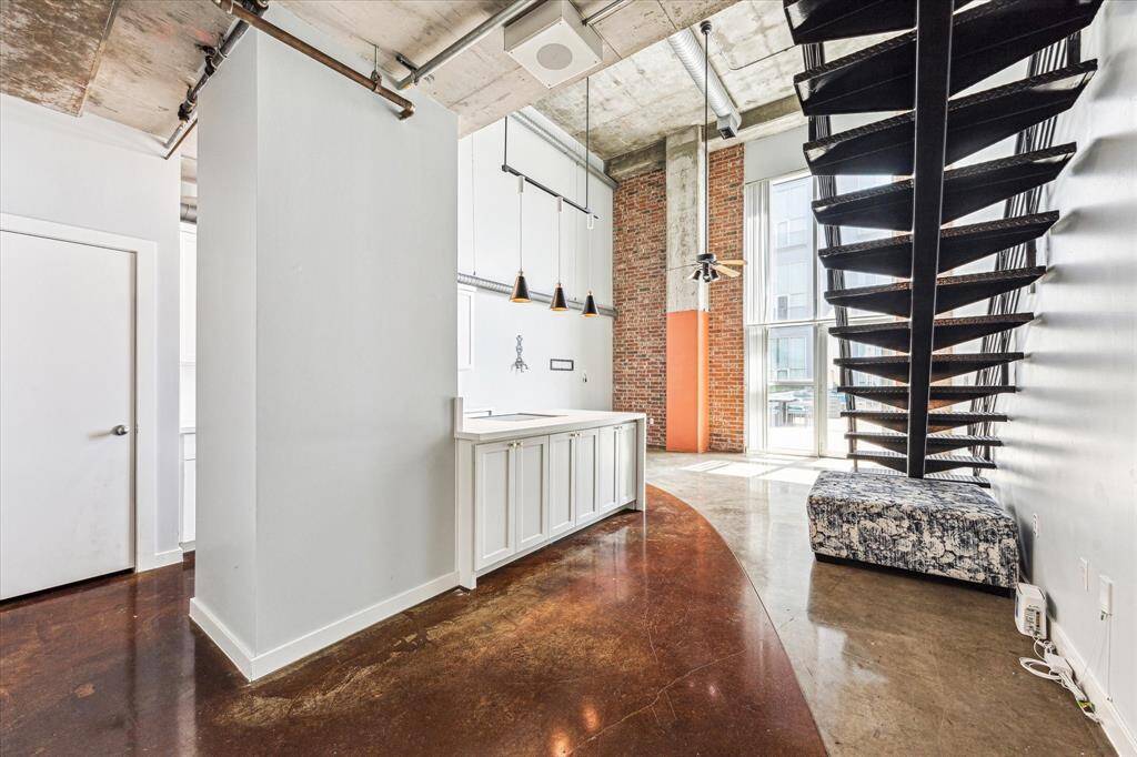 This modern loft features an open layout with high ceilings, exposed concrete, and a striking metal staircase. The space is brightened by large windows and includes a sleek kitchen area with pendant lighting. Ideal for urban living with a stylish industrial vibe.