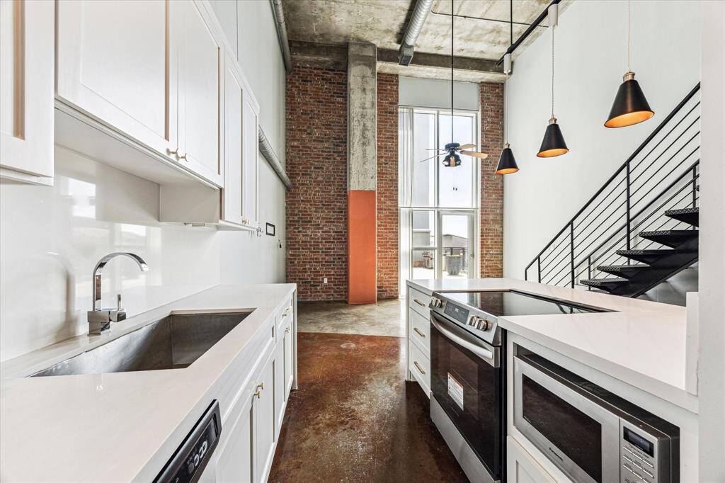 This modern kitchen features high ceilings, exposed brick, and industrial accents. It includes sleek appliances, white cabinetry, and pendant lighting, leading to an open loft with a metal staircase.