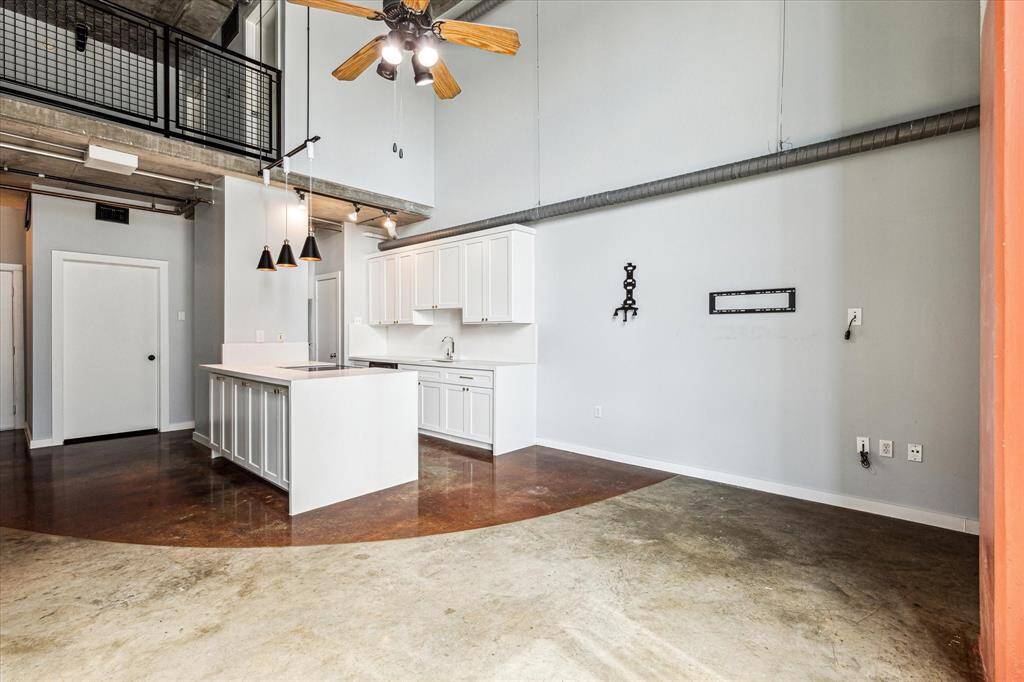 Modern loft-style home with high ceilings and an open layout. The kitchen features white cabinetry and a central island. The space includes industrial elements like exposed ducts and a ceiling fan, with polished concrete floors adding a sleek touch.