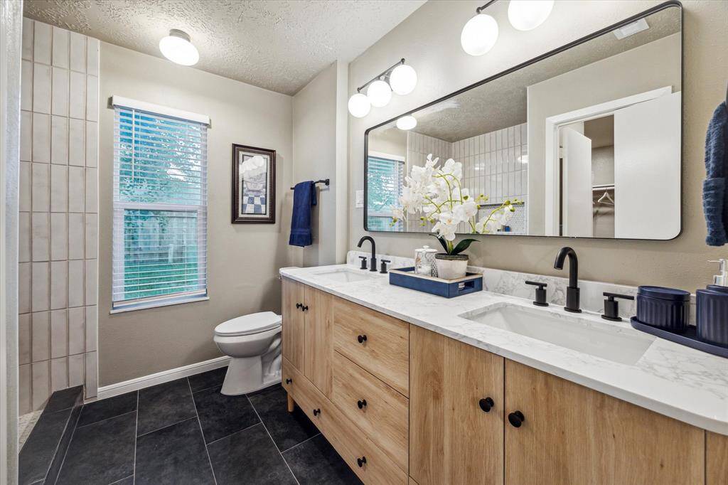 The primary ensuite bath features a sleek double vanity with wood cabinetry, quartz countertops, and matte black fixtures. A large framed mirror and modern lighting enhance the bright, stylish design. The large walk-in shower, paired with elegant tile flooring, creates a spa-like retreat.