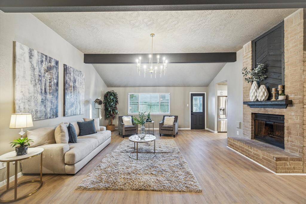 This spacious living room features vaulted ceilings, exposed beams, and a stunning chandelier. A brick fireplace with a modern black accent adds warmth and character. The open layout, natural light, and stylish finishes make it perfect for entertaining.