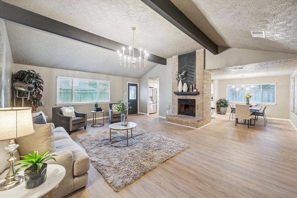 This open-concept living and dining space features high ceilings, exposed beams, and abundant natural light. The centerpiece brick fireplace adds warmth, while modern lighting enhances the stylish design. With seamless flow to the kitchen and dining area, it’s perfect for gatherings.