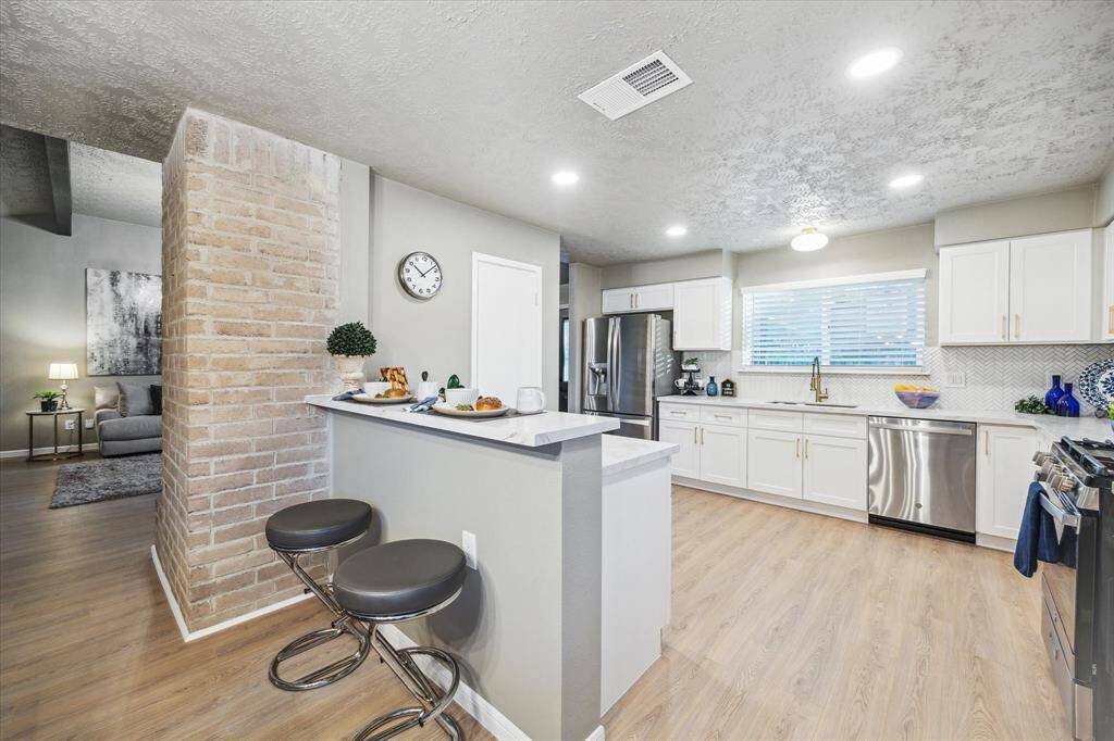 This beautifully updated kitchen features sleek white cabinetry, stainless steel appliances, and a spacious layout. A stylish breakfast bar with modern seating overlooks the living area, creating a perfect gathering spot. The warm wood flooring and brick accent wall add character and charm.
