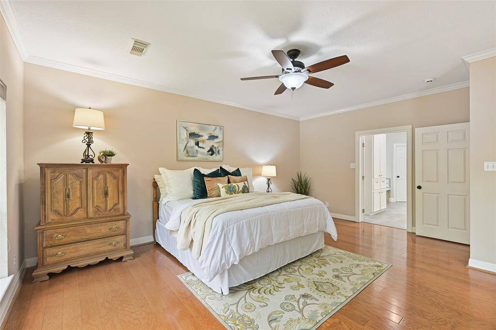 Escape to the first-floor owners' retreat that showcases more wood floors, a lighted ceiling fan, neutral paint trimmed with crown molding, an expansive rear-facing window with pleated shades, and French doors allowing a private exit to the covered patio area.