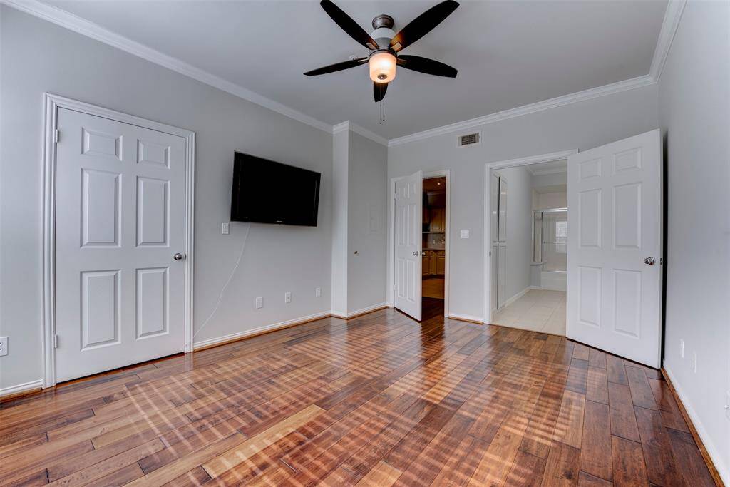 Alternate view of the primary bedroom and its hardwood floors, a ceiling fan with light kit, a wall-mounted TV - all lined with crown molding. Door to the right leads to an en-suite bathroom.