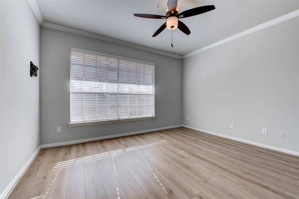 A sizable secondary bedroom shows a bright room with light wood flooring, neutral  walls, and a ceiling fan with light kit.