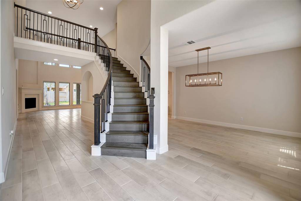 This is a spacious, modern interior featuring a grand staircase with dark steps and a wrought-iron railing, leading to an upper level with an open gallery. The room has light-colored tile flooring, neutral wall tones, and ample natural light from large windows and a glass door that provide a view to the outside. A sleek rectangular chandelier hangs above, adding a touch of elegance to the space.