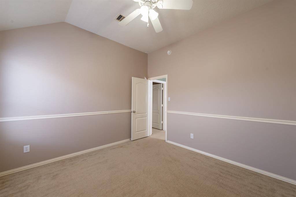 This room features neutral walls with a chair rail, a vaulted ceiling with a ceiling fan, and plush carpet. There's a white door leading to a hallway, adding to its cozy and inviting feel.