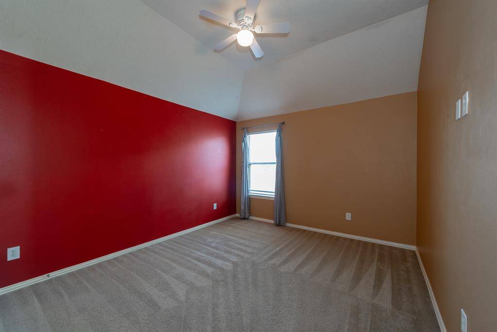 This room features a high ceiling with a ceiling fan, a bold red accent wall, and neutral carpet. A window with curtains allows natural light, complementing the warm wall tones.