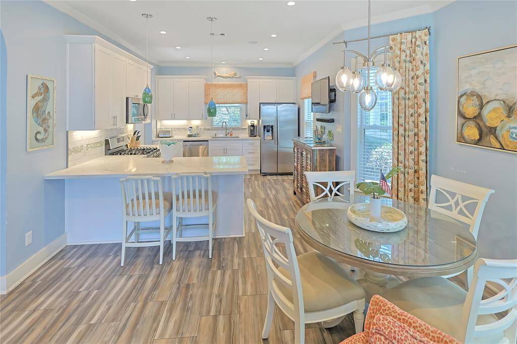 This bright kitchen and dining area features modern appliances, a large island with seating, and stylish pendant lighting. The space is accented with wood-like flooring, and a cozy dining nook, creating a welcoming and functional living area.