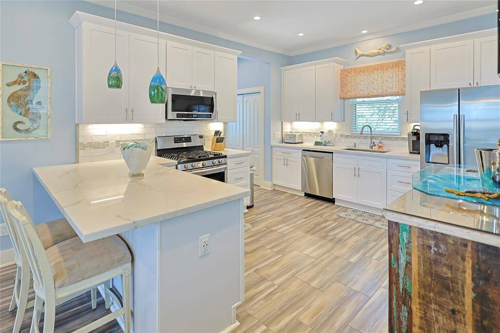 This beautiful kitchen features a modern design with white cabinetry and stainless steel appliances. It has a spacious island with seating, colorful pendant lighting, and a stylish wood-like tile floor. The room is bright, with ample natural light and a decorative coastal theme.