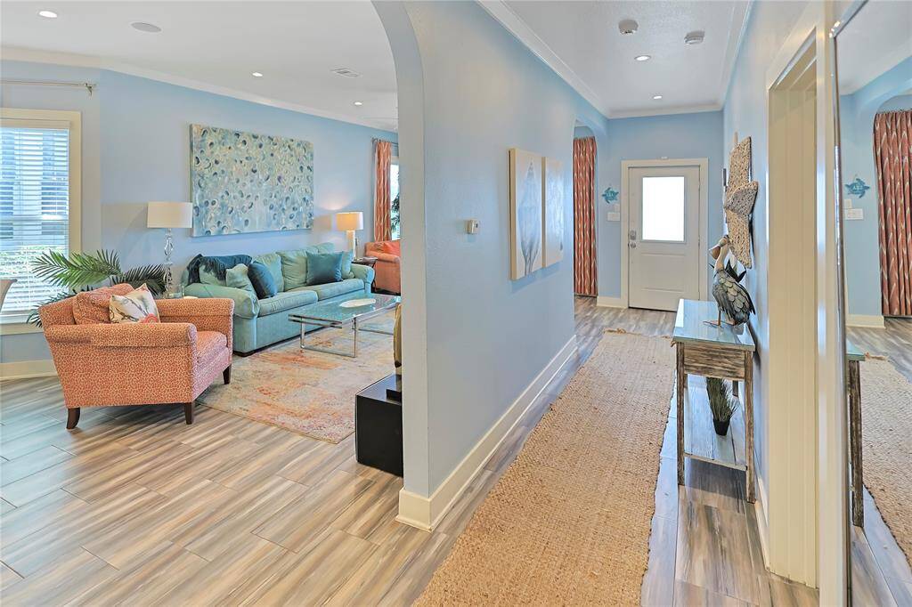 This inviting home features a bright, open living area with light blue walls, cozy seating, and modern decor. The hallway leads to a welcoming front door, with stylish wood-look flooring throughout, creating a warm and contemporary atmosphere.
