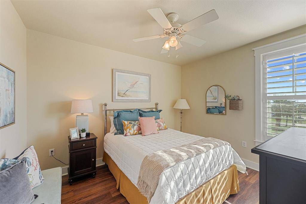 This sunlit guest bedroom offers a cozy place to unwind with coastal-inspired decor, wood-style flooring, and beautiful views through plantation shutters.