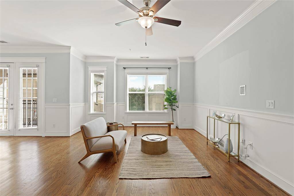 Spacious living room featuring hardwood floors, elegant wainscoting, and crown molding. Natural light streams in through large windows, complemented by a ceiling fan.