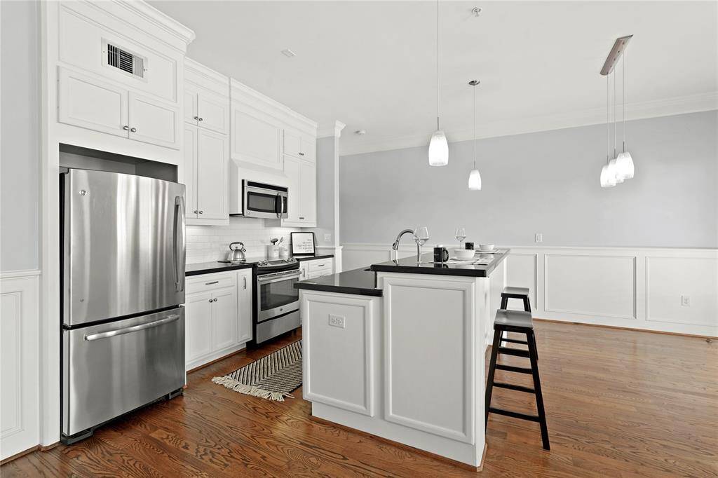 Spacious and modern kitchen with white cabinetry, stainless steel appliances, a central island with seating, and hardwood flooring that flows into an open-plan living area with ample natural light.