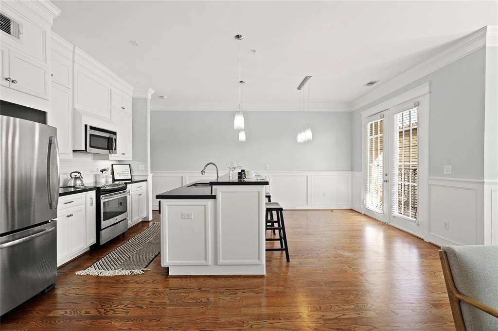 Spacious and modern kitchen with white cabinetry, stainless steel appliances, a central island with seating, and hardwood flooring that flows into an open-plan living area with ample natural light.