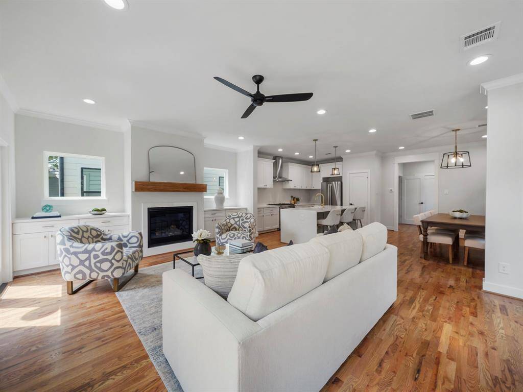 Bright and inviting living room featuring rich hardwood floors, a cozy fireplace, and large windows that fill the space with natural light. The open floor plan seamlessly connects to the kitchen and dining area, creating a perfect setting for entertaining. A door leads to the back porch, offering a smooth transition to outdoor living.