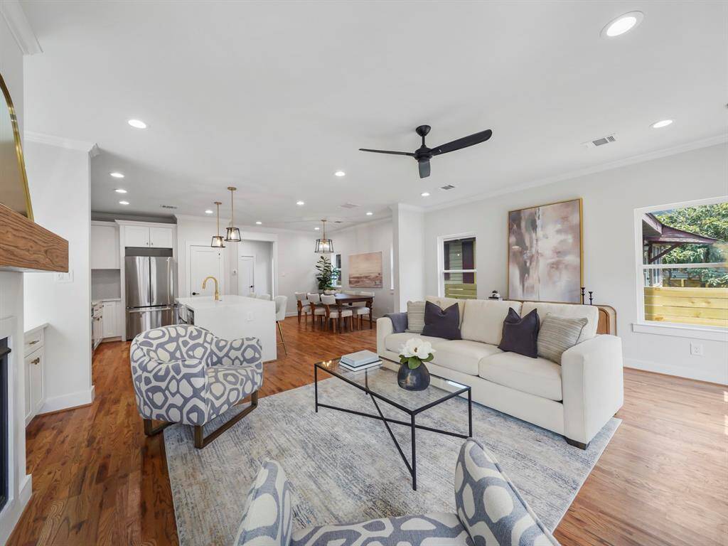 Bright and inviting living room featuring rich hardwood floors, a cozy fireplace, and large windows that fill the space with natural light. The open floor plan seamlessly connects to the kitchen and dining area, creating a perfect setting for entertaining. A door leads to the back porch, offering a smooth transition to outdoor living.