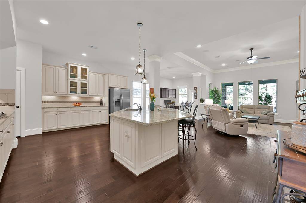 Open-concept kitchen and living area with a spacious layout. It features a large island with seating, elegant lighting, and ample cabinetry. The living space includes plenty of space for comfortable seating and large windows, offering a bright and inviting atmosphere.