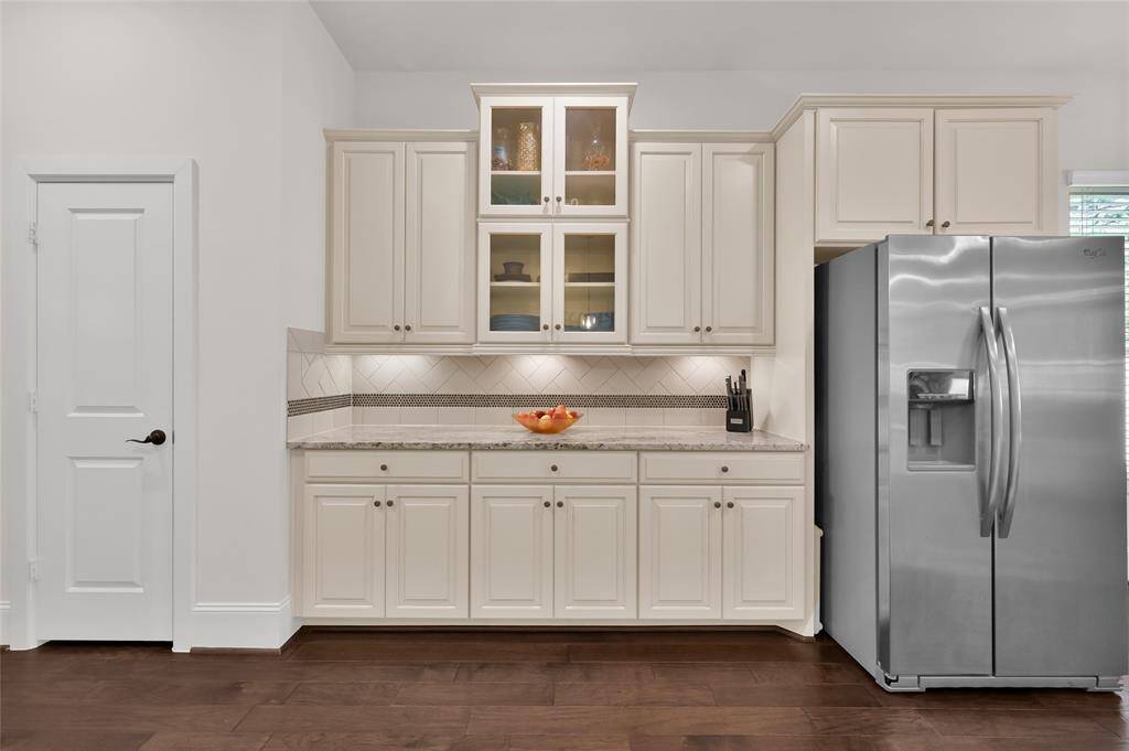 This kitchen features elegant cabinetry with glass-front accents, a modern stainless steel refrigerator, and a sleek granite countertop. The backsplash adds a stylish touch, complemented by dark wood flooring for a warm, sophisticated look.