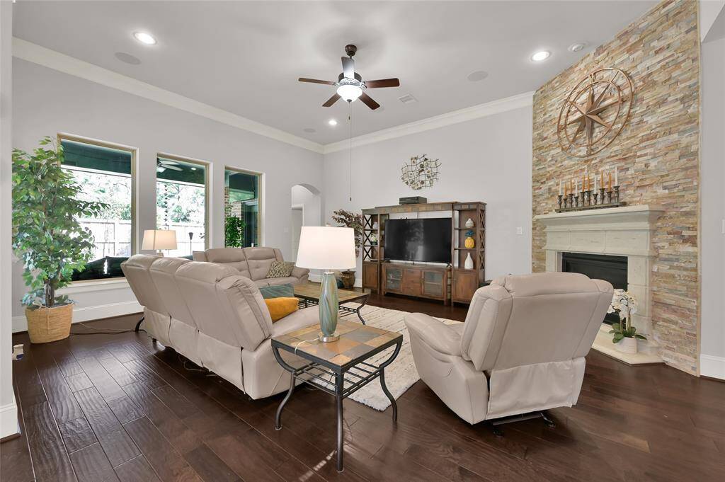 This spacious living room features elegant hardwood flooring, a cozy stone fireplace, and large windows for natural light. It includes comfortable seating, a stylish entertainment center, and tasteful decor, creating a warm and inviting atmosphere.
