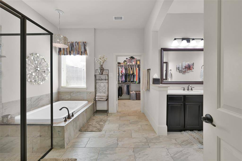 This spacious primary bathroom features a large soaking tub with a modern glass shower enclosure. The room is well-lit with a stylish light fixture and natural light from the window. It includes a double vanity with dark cabinetry and a walk-in closet, offering ample storage and convenience.