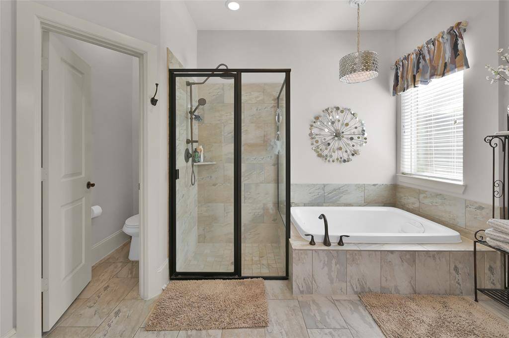 This bathroom features a spacious walk-in shower with glass doors, a large bathtub adjacent to a window with natural light, and a separate toilet area. The neutral tones and modern fixtures add elegance and comfort.