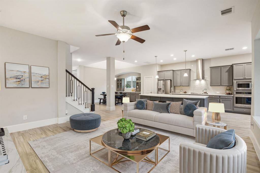 This spacious open-concept living area features a modern kitchen with stainless steel appliances and a large island, seamlessly connecting to a stylish living room with contemporary furnishings and a ceiling fan. Natural light fills the space, enhancing the warm, neutral tones and elegant design.