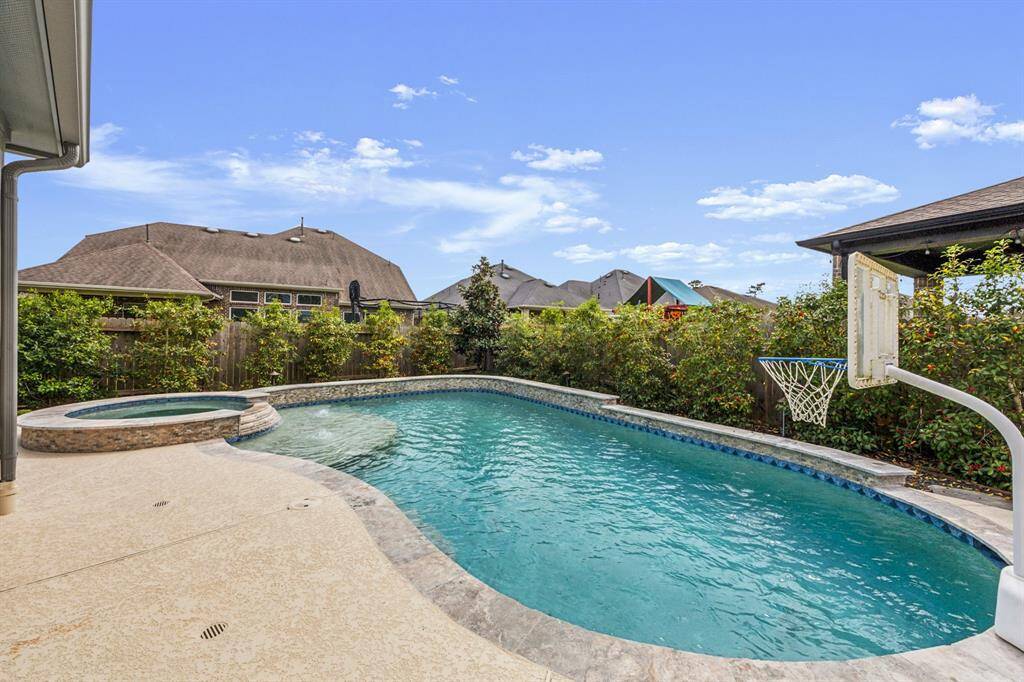 This backyard features a spacious swimming pool featuring a built-in spa. The pool area is surrounded by lush greenery and includes a basketball hoop, offering a private and recreational outdoor space perfect for relaxation and entertainment.